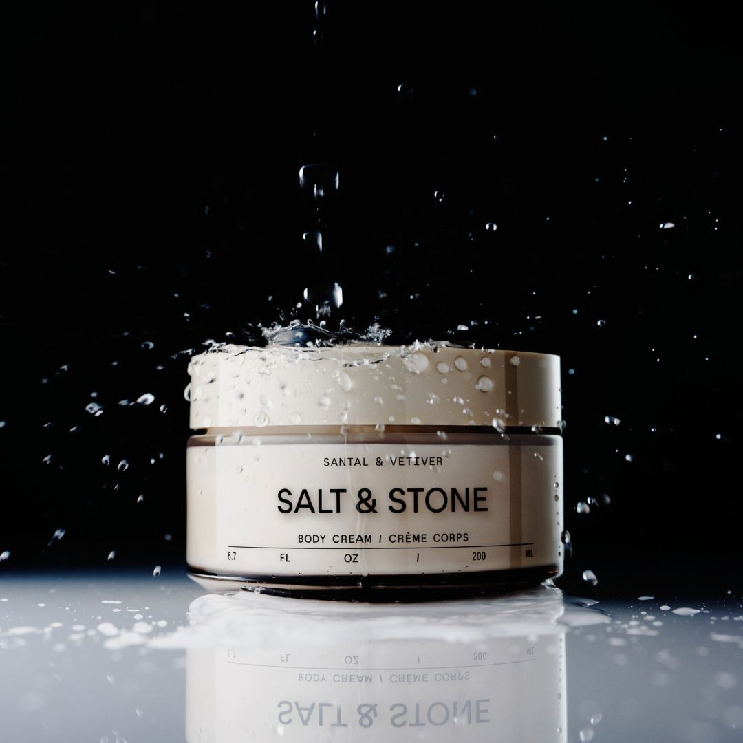 A jar of Santal & Veijver Salt & Stone body cream sits on a reflective surface with water droplets splashing and dripping off it against a black background.