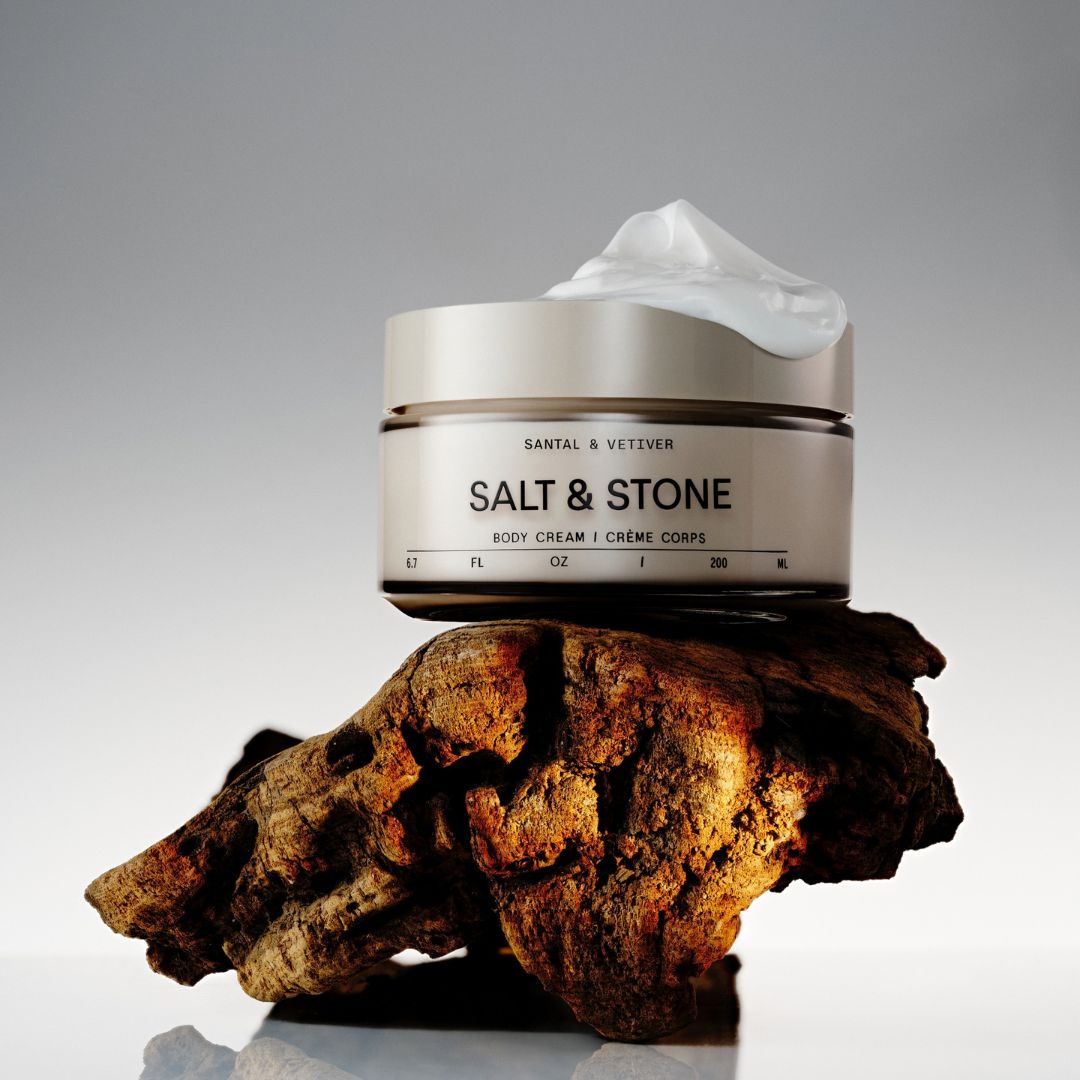 A jar of Santal & Vettver Salt & Stone body cream sits on a textured piece of driftwood against a neutral gray background. The cream is visible as a dollop on top of the jar.