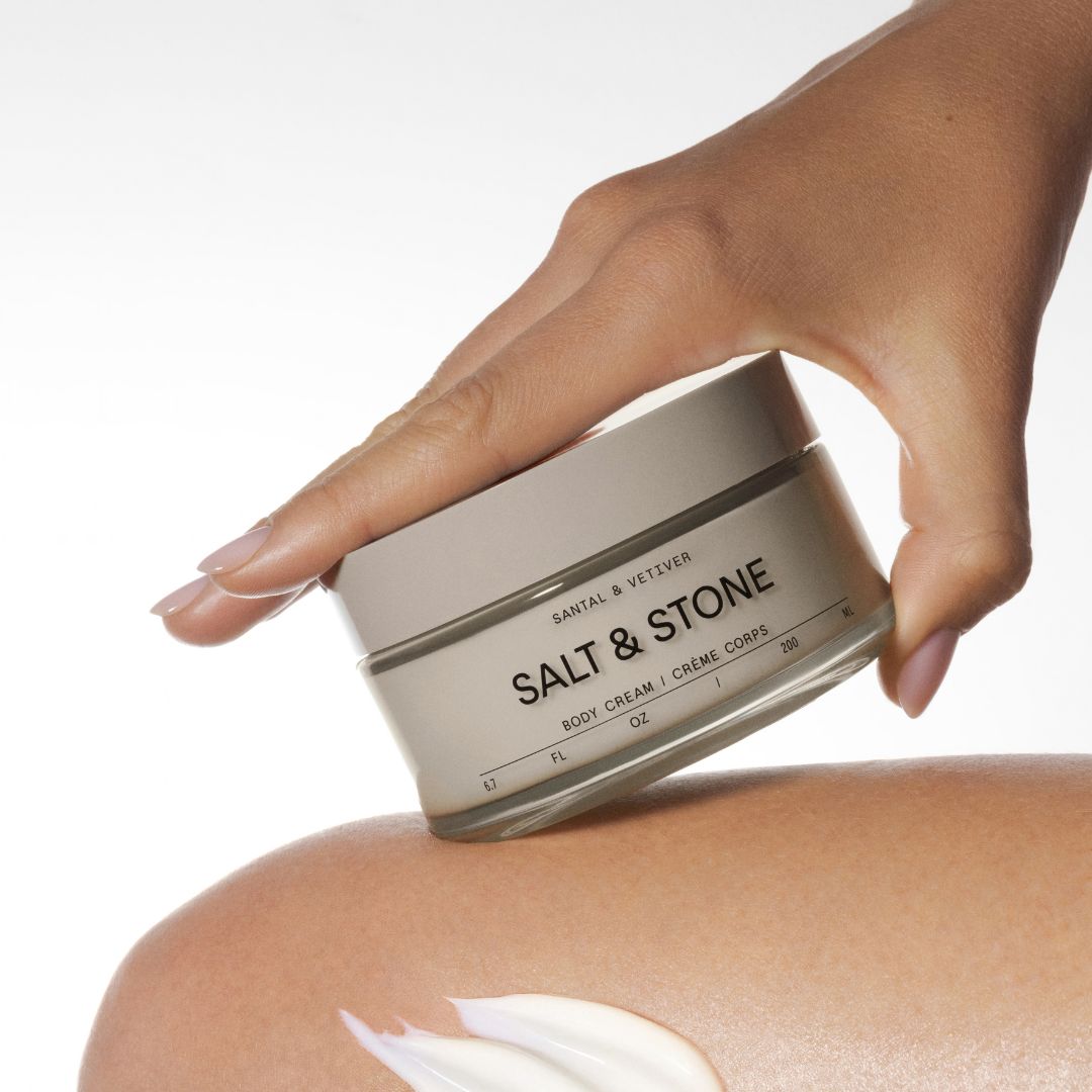 A hand holds a silver jar of Salt & Stone body cream against a clean white background, with a dollop of the white cream visible on skin below.